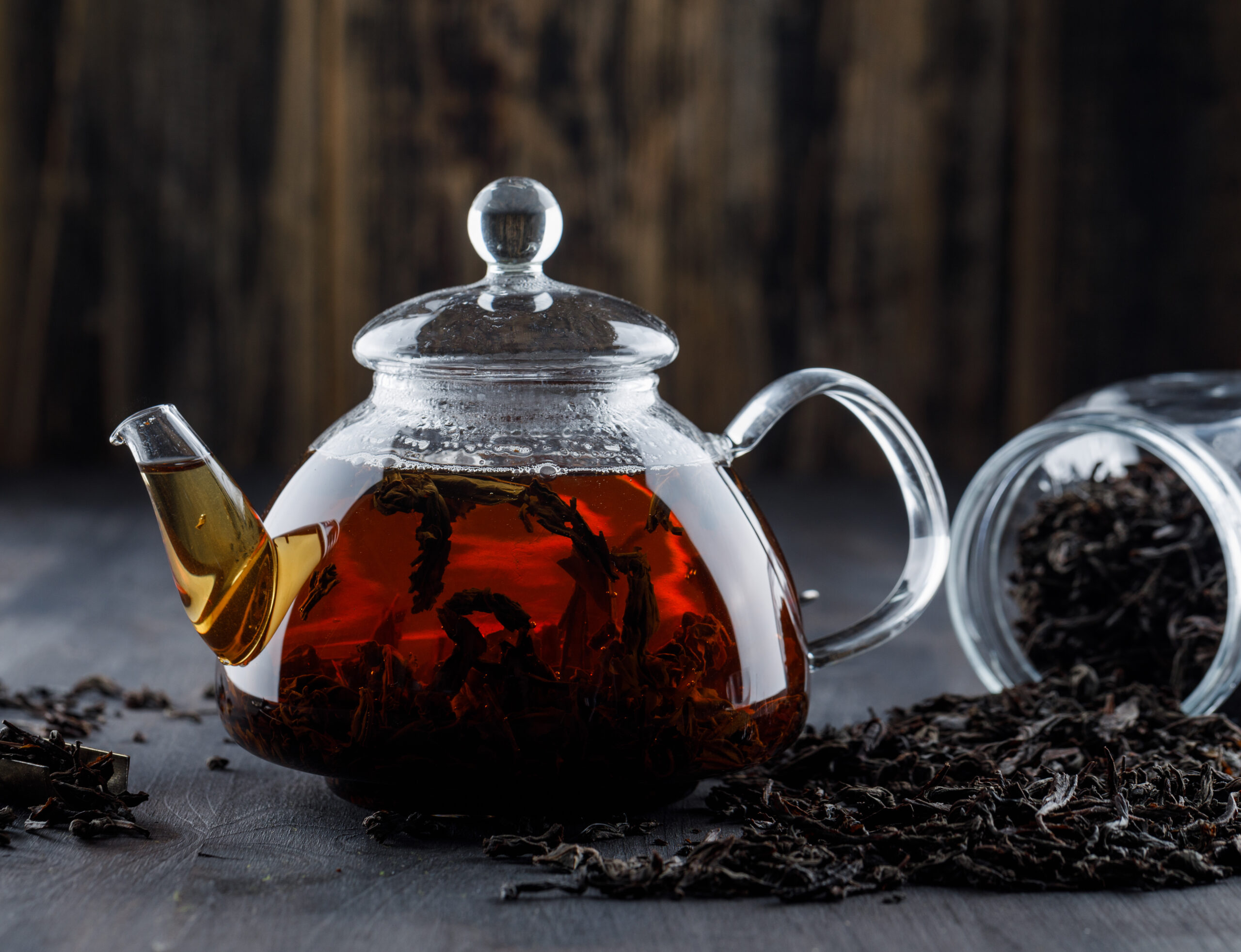 Glass teapot filled with brewed black tea and loose leaf tea on rustic wooden surface